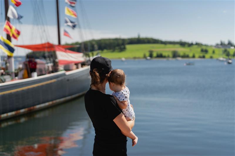 A woman holding a baby in front of a sailboat in Halifax, Canada.