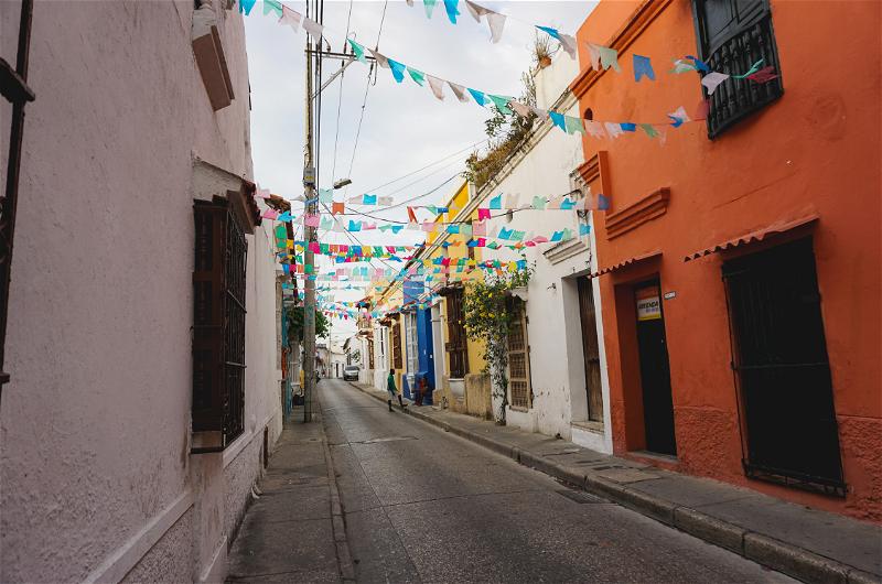 Small multicolored flags hang across a small quiet street with white and orange buildings