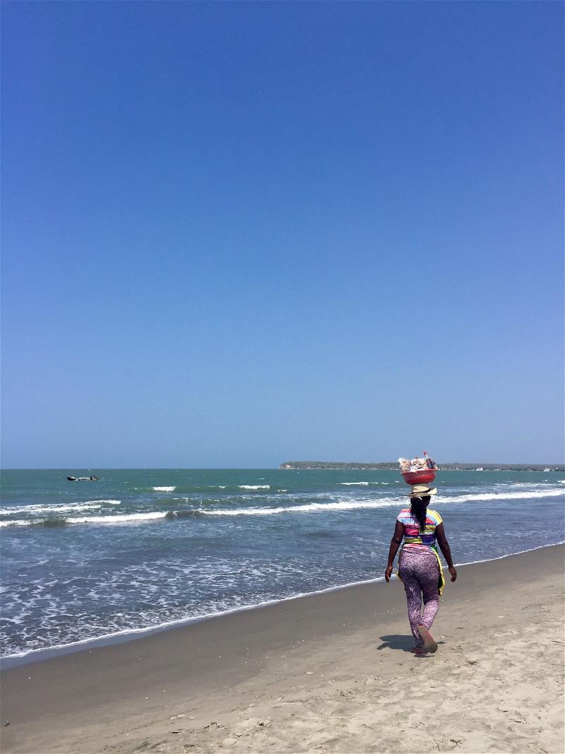 A woman walking on the beach with a basket on her head.