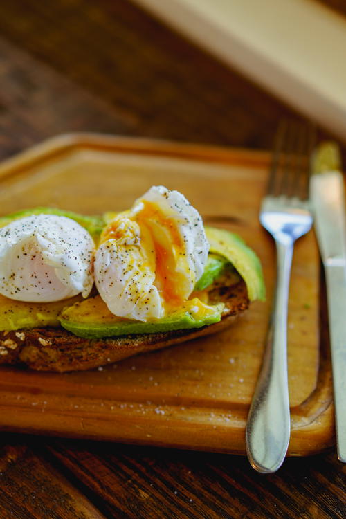 A toast with eggs and avocado on a wooden cutting board.