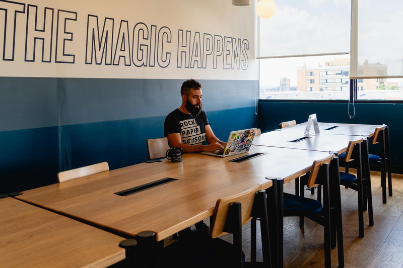 A man sits at a table with a laptop in front of a wall that says the magic happens.