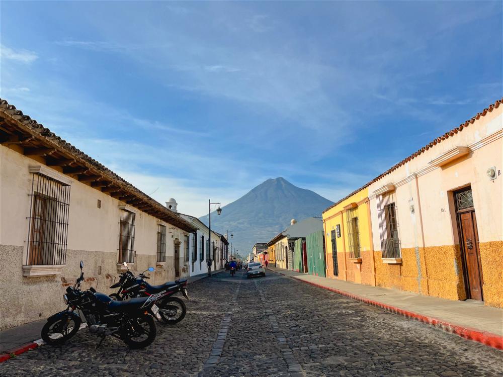 A narrow street in Guatemala with motorcycles parked in front of a volcano.