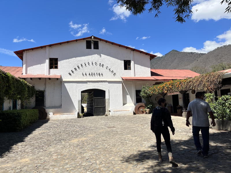 Two individuals pass by a white building in Guatemala, as a majestic mountain looms in the background.