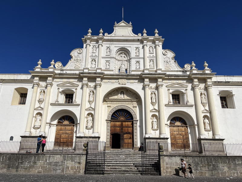 The front of a white church in Guatemala with people walking in front of it.