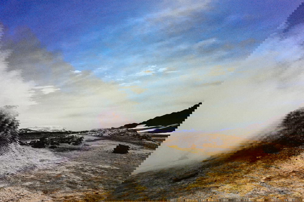 A man sitting on a sand dune next to a steaming geyser in Iceland.