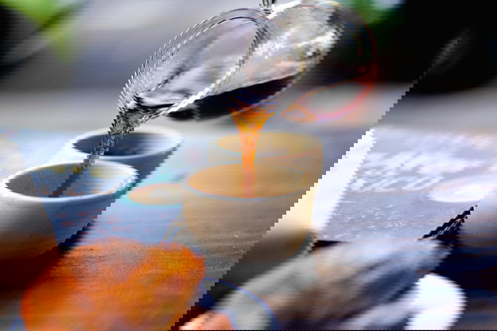 Pouring a stream of black coffee from a glass carafe on a wooden table into a small white cup