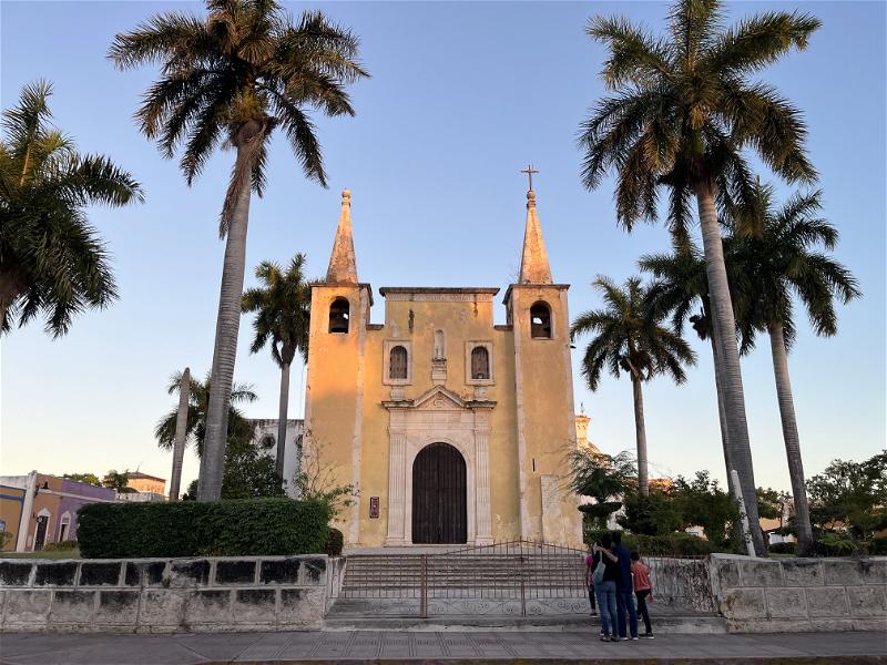 Two people standing in front of a church in Merida, Mexico, surrounded by palm trees.