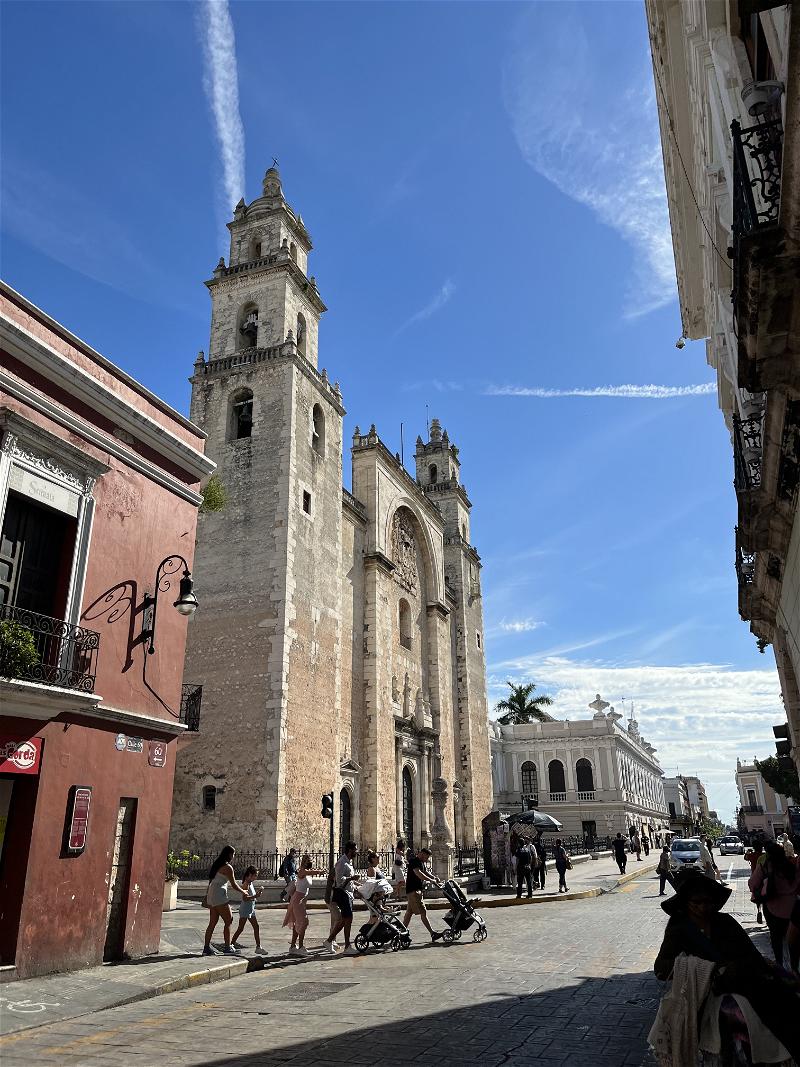 People walking down a street in Merida, Mexico with a church tower in the background.