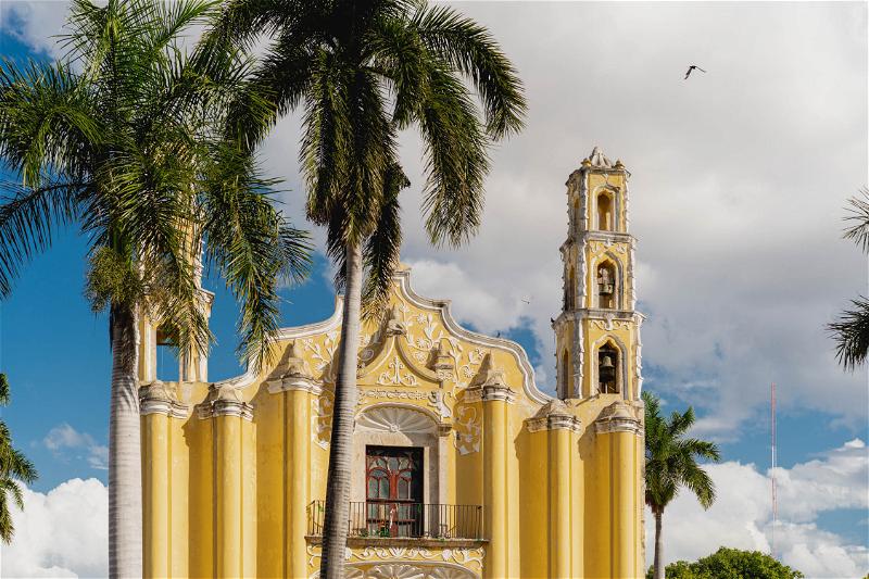 A yellow church in Merida, Mexico with palm trees in front of it.
