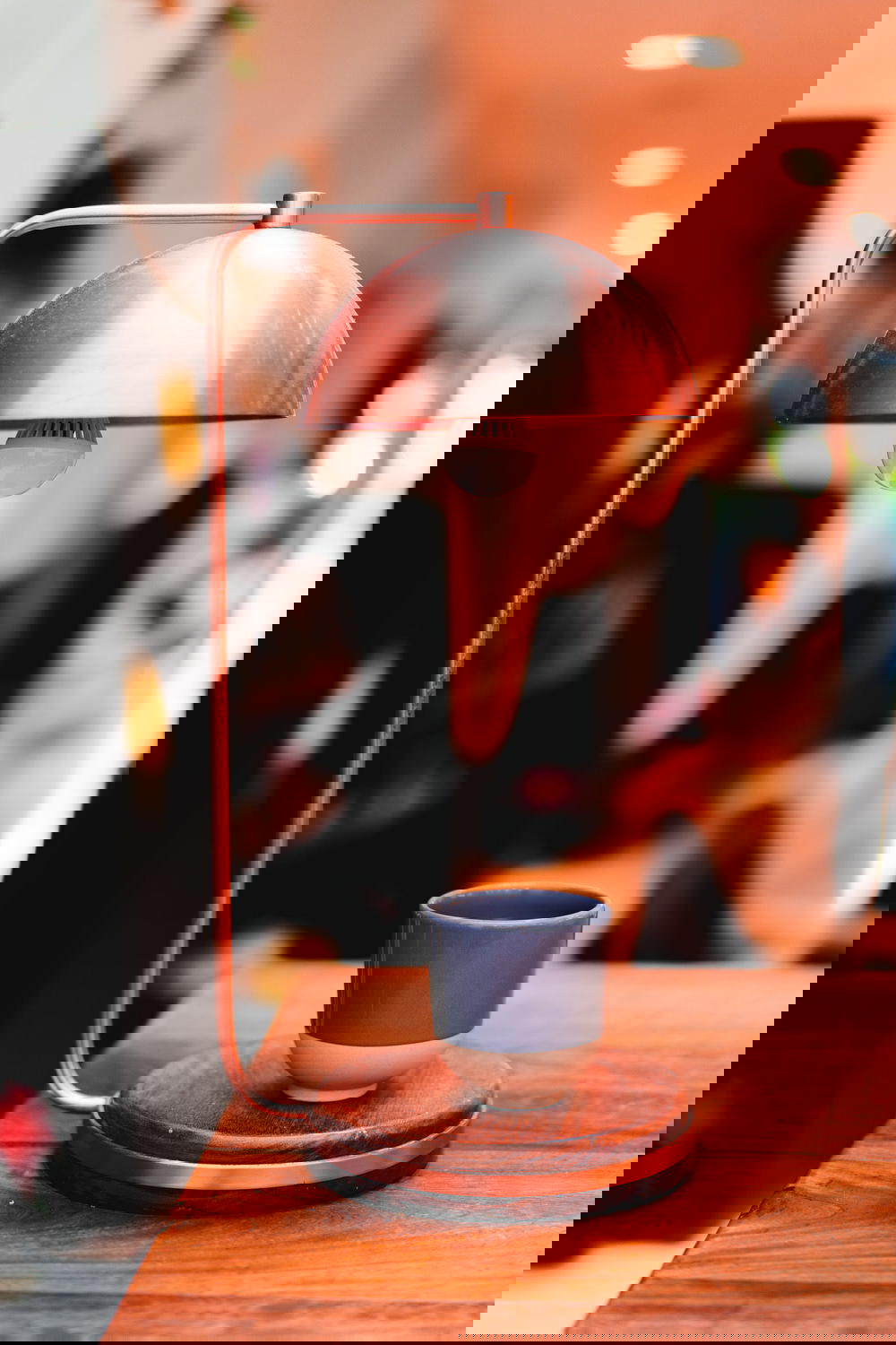 Rose colored brushed metal lamp with a wooden base and a blue cup of coffee on a wooden table