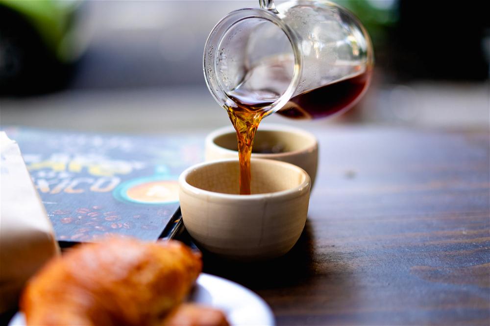 Pouring a stream of black coffee from a glass carafe on a wooden table into a small white cup