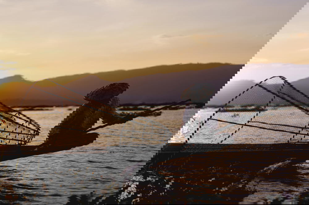 Burmese dancing fishermen sitting on the edge of a small fishing boat at sunset on Inle Lake