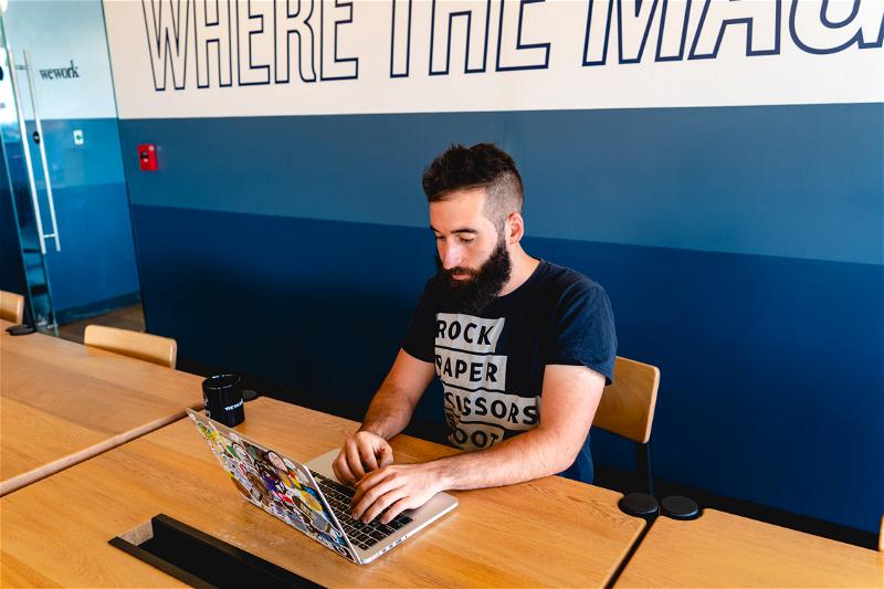 Man working remotely from a WeWork hot desk at a communal wooden table against a blue wall