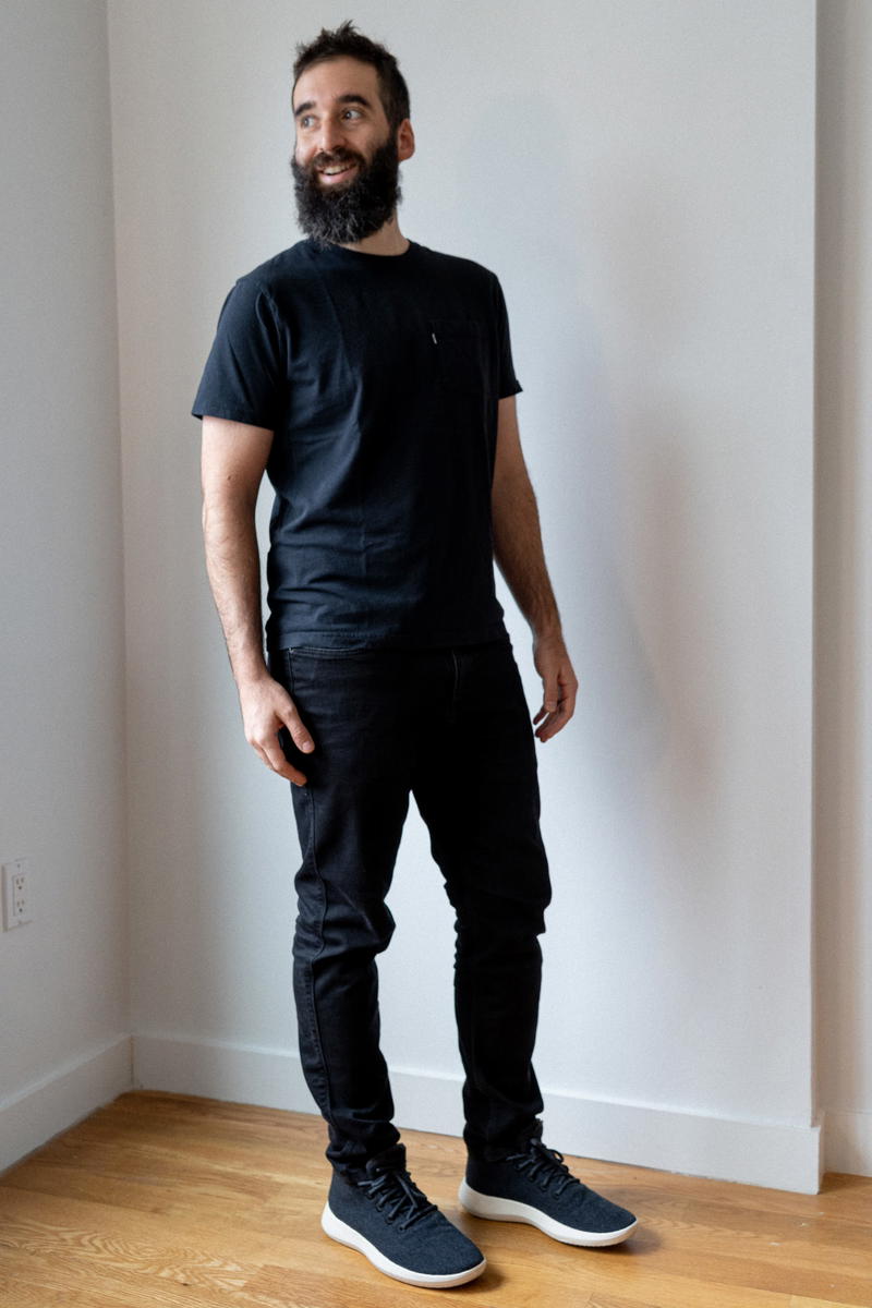 A bearded man standing in a room wearing black t - shirts and black jeans.