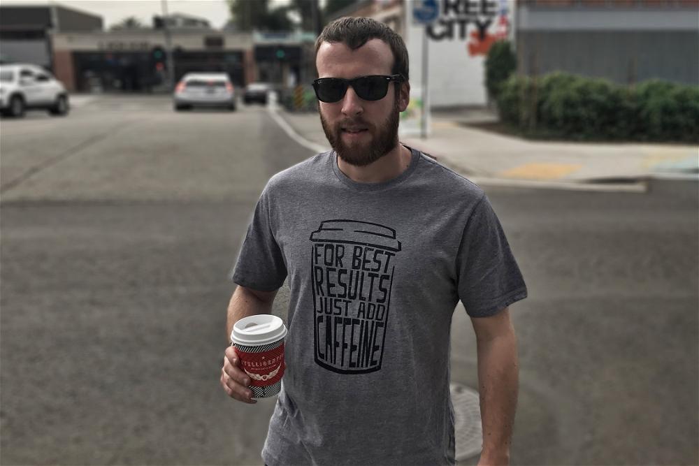 A bearded man wearing a grey t - shirt holding a cup of coffee.