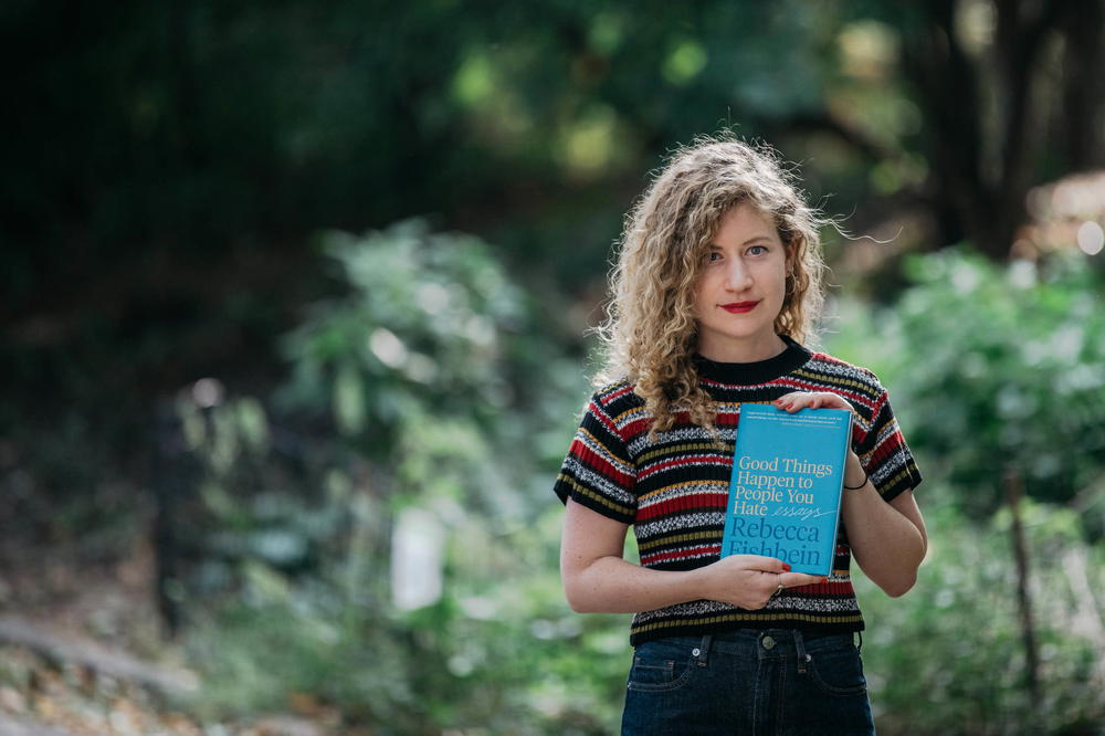 A young woman holding a blue book in front of trees.