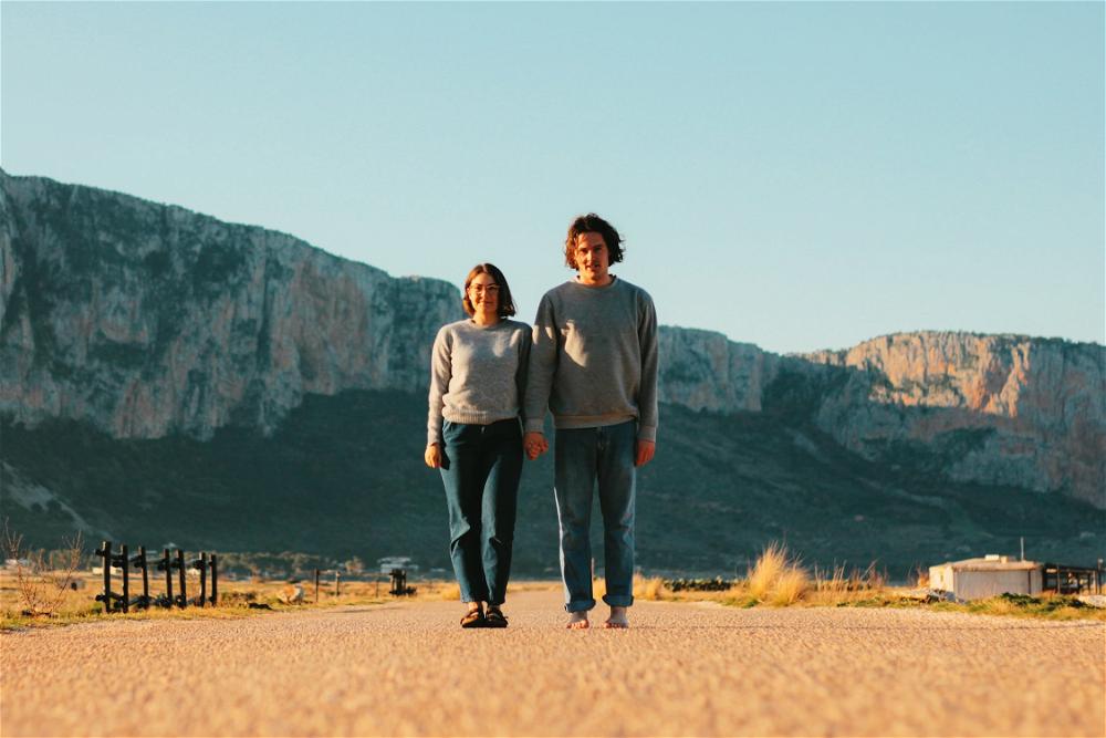 Two people standing on a road with mountains in the background.