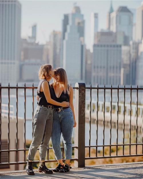 Two women hugging in front of a fence with a city skyline in the background.