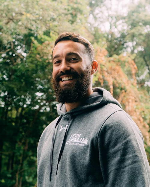 A man with a beard smiling in front of trees.