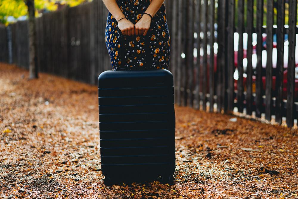 A woman holding a black suitcase in front of a fence.