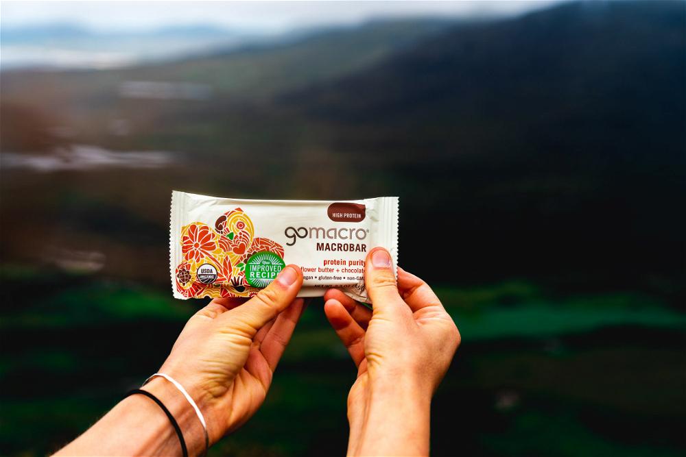 A person holding a bar of granola in front of a mountain.