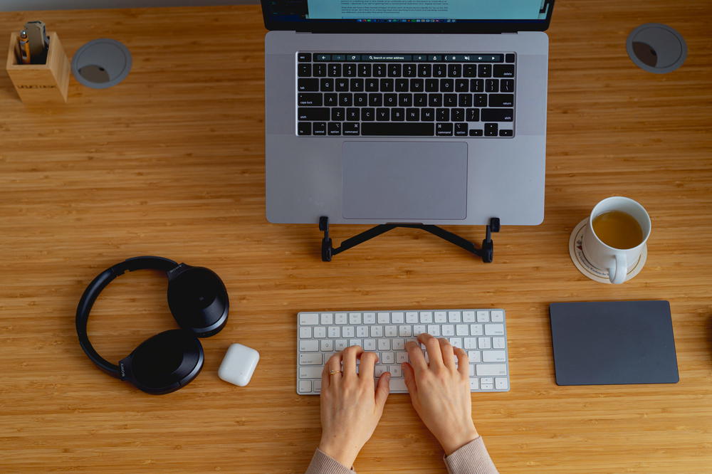 Minimalist home office setup of black headphones, an apple magic keyboard, silver MacBook Pro on a laptop stand and cup of tea