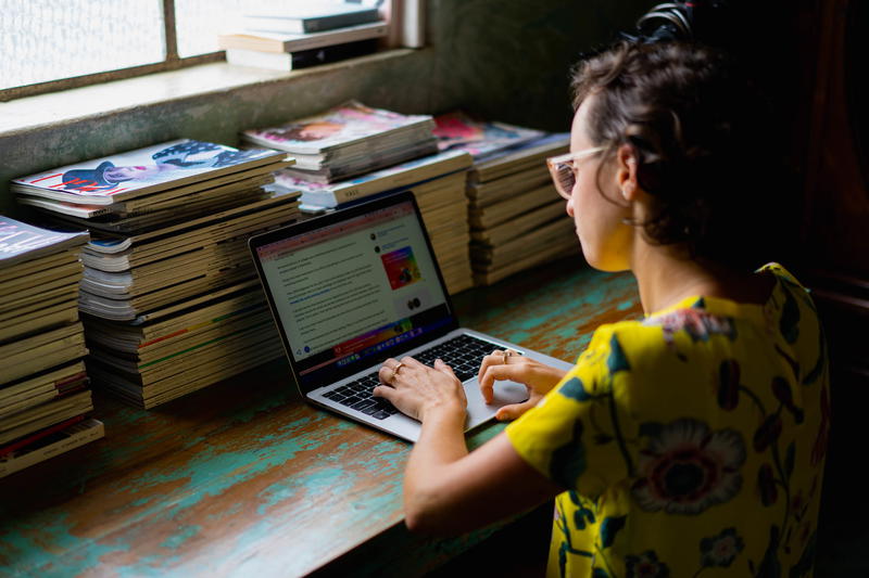 A woman working on a laptop in front of a pile of books.