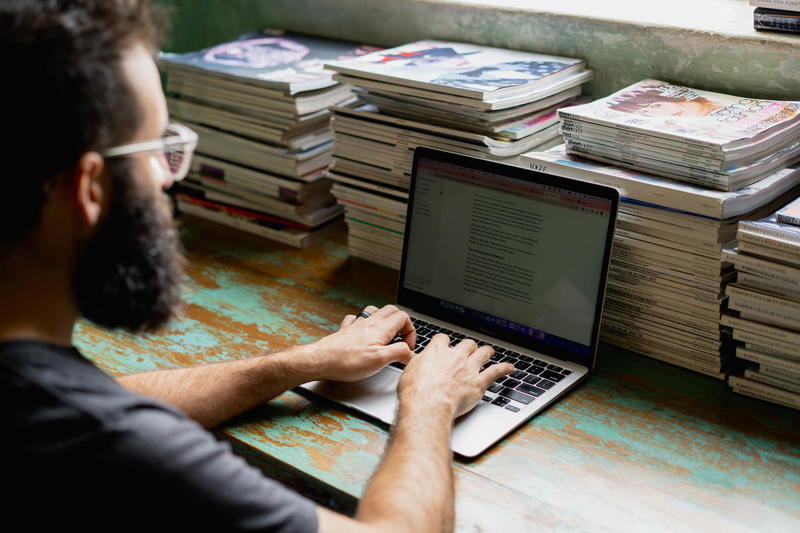 A man typing on a laptop in front of a pile of books.
