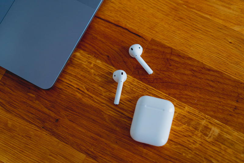 Two AirPods and an AirPod case on a wooden table next to a silver MacBook in a home office