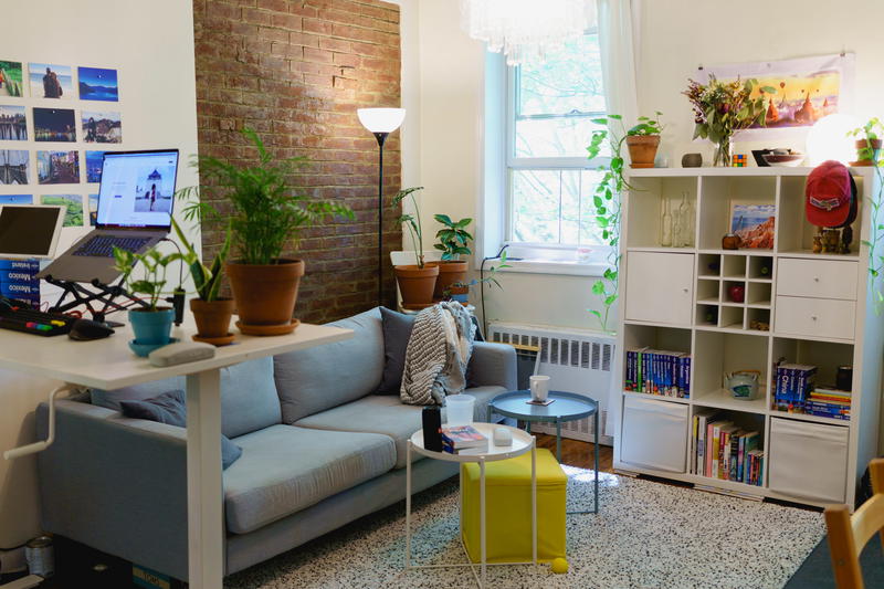 Standing desk setup in a home office design in a small apartment with a couch, ottoman, bookshelf and exposed brick wall