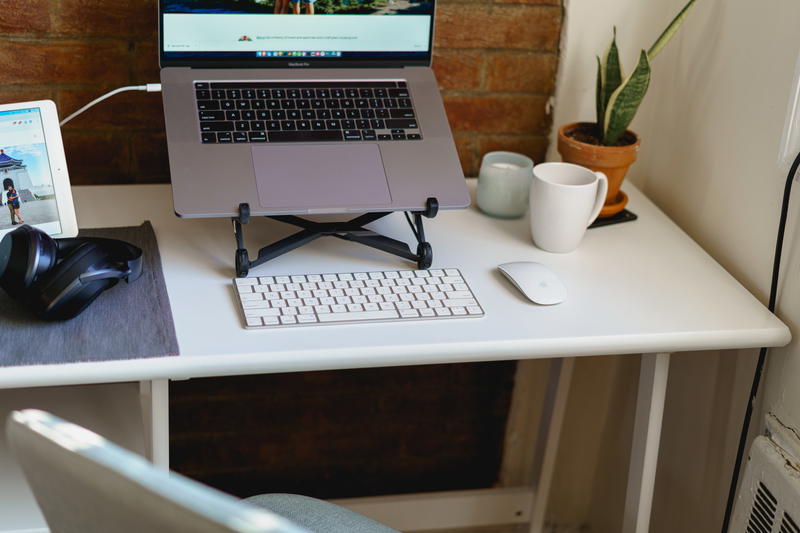 Small slim white writing desk for home office set up with laptop, wireless keyboard, light green desk chair and white wireless mouse against a brick wall