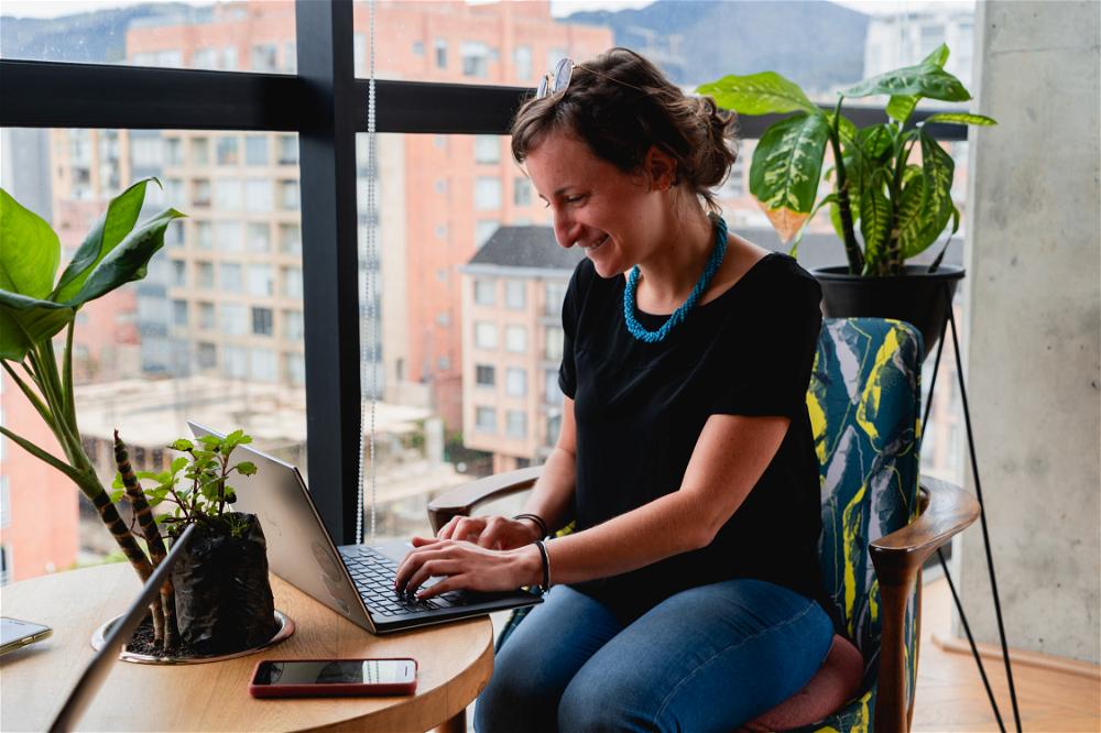 Woman working remotely from a laptop at a round wooden table in a coworking space with plants