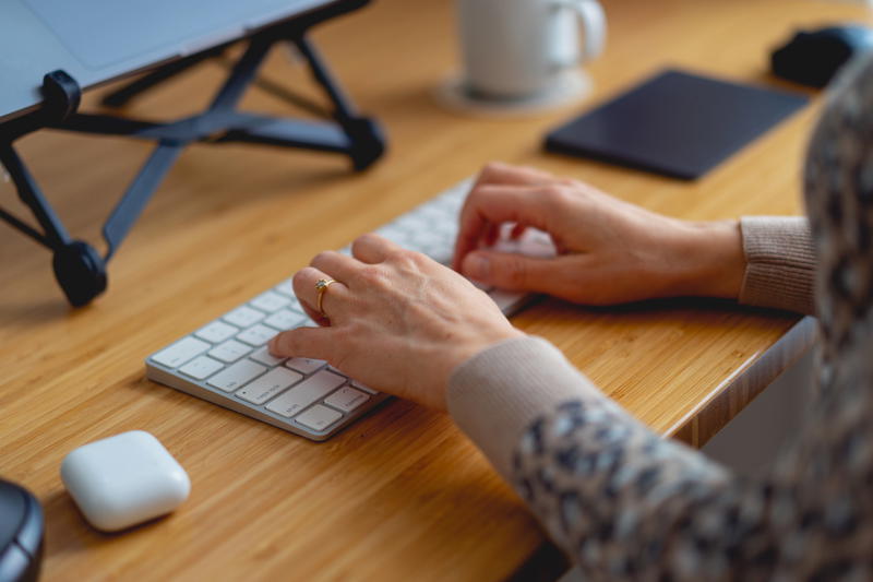 Woman typing on a white external Apple Magic keyboard on a bamboo desk in a home office with a laptop stand and AirPod case