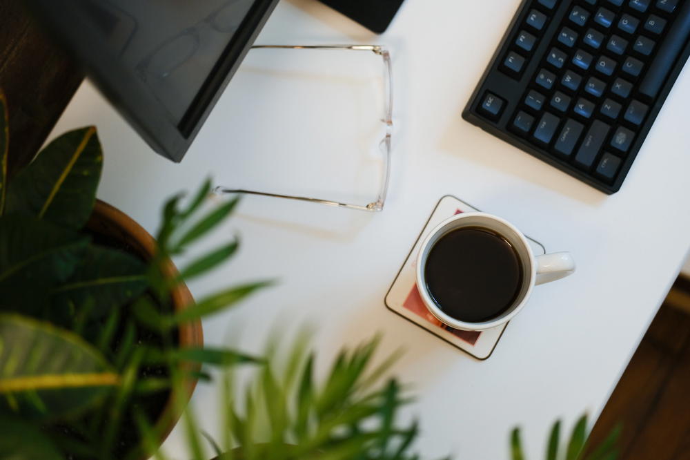 Flat lay of a home office setup with clear glasses, a white cup full of coffee, leafy green plants and a black keyboard