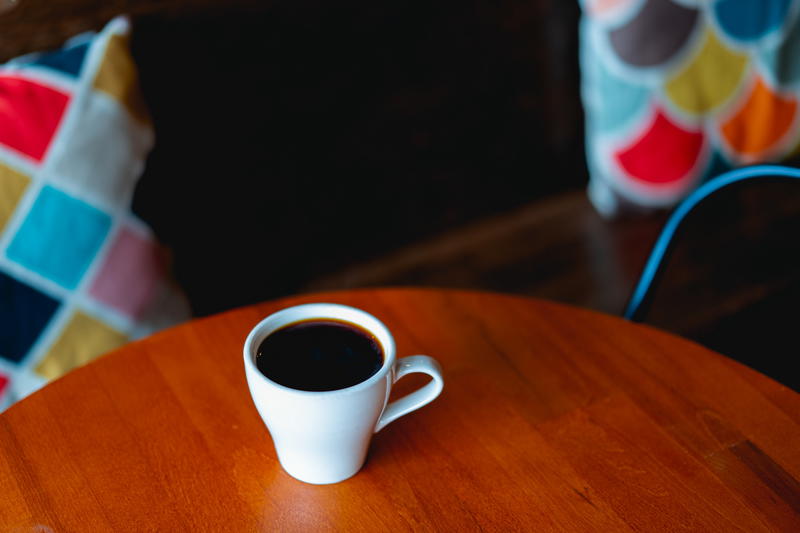 White coffee cup full of coffee on a wooden circular table with patterned pillows on a bench