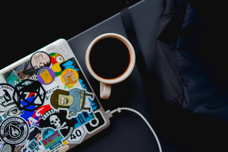 White coffee mug full of coffee on a black surface next to a black backpack and a laptop plugged in