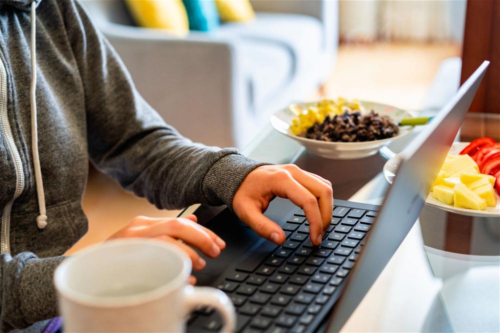 Woman wearing a gray hoodie typing at a laptop with a white teacup and two bowls of healthy lunch food working at home