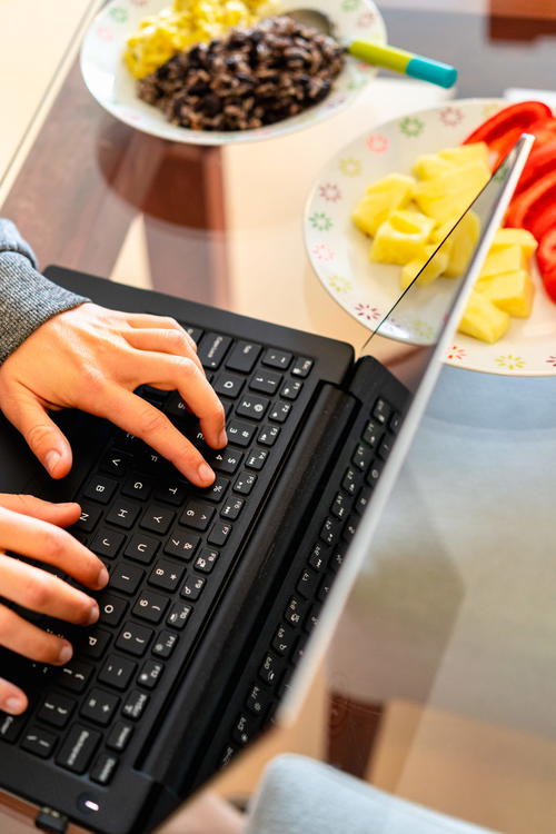 Woman typing on a black 13 inch laptop on a glass table next to plates of food