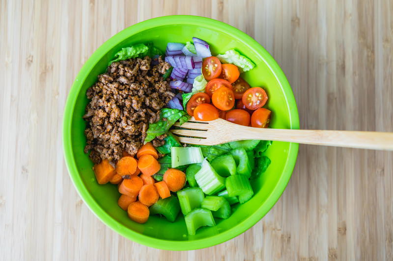 Green bowl of ground beef, chopped carrots, chopped celery, chopped cherry tomatoes and chopped red onion with a bamboo fork