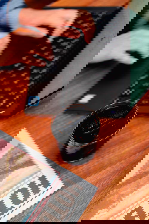 Delicate glass full of cold brew iced coffee next to a black laptop with a girl typing on the keyboard