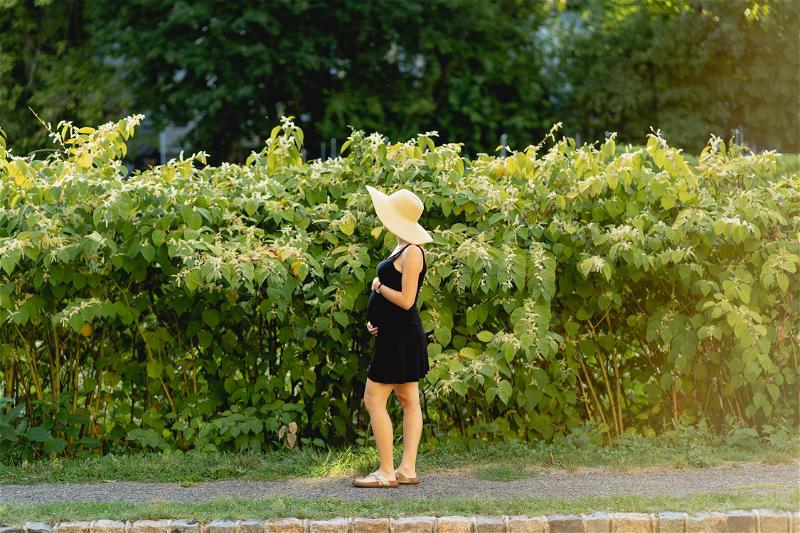 A pregnant woman in a hat standing next to a hedge.