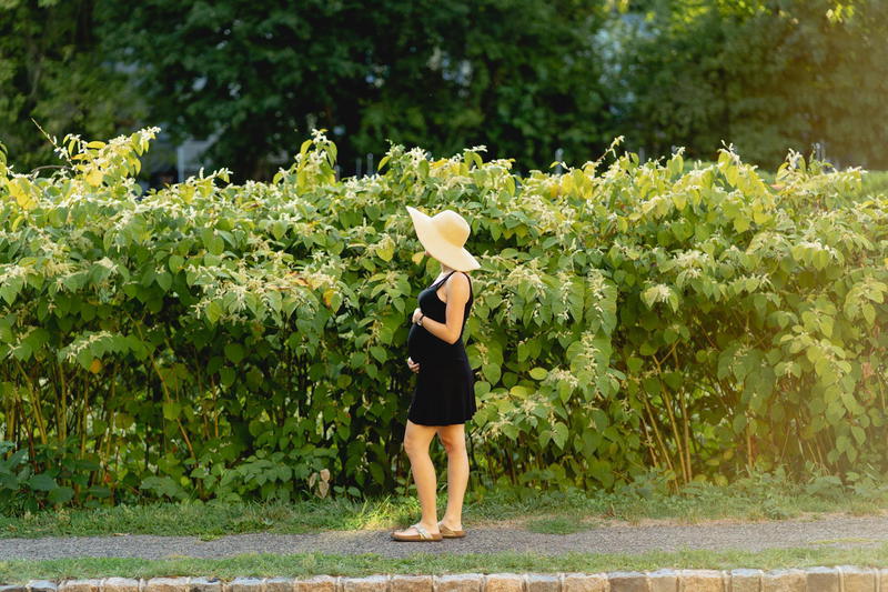 A pregnant woman in a hat standing next to a hedge.