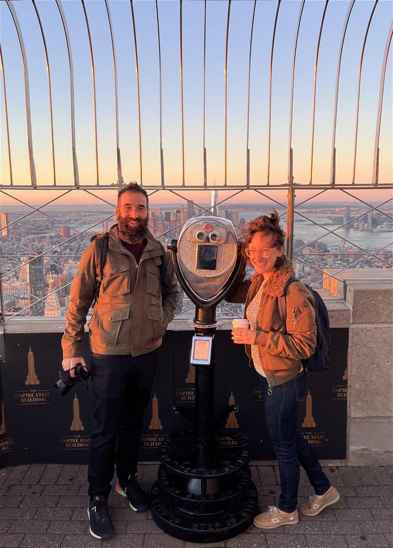 Two people posing in front of a telescope on top of the empire state building.