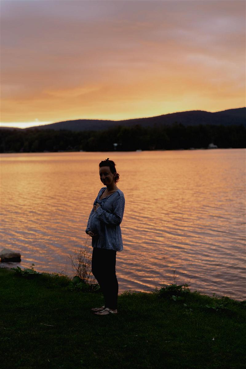 A pregnant woman standing in front of a lake at sunset.