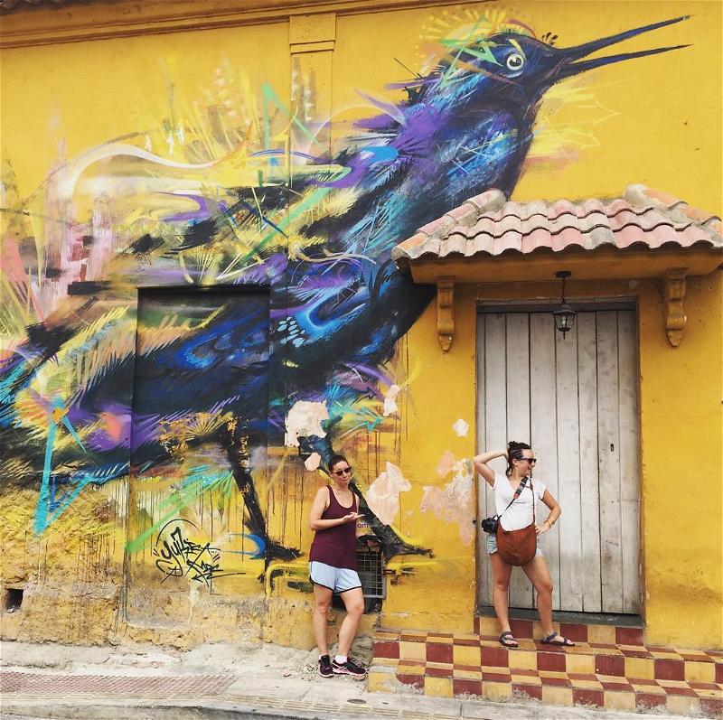 Two women standing in front of a yellow building with a bird painted on it.