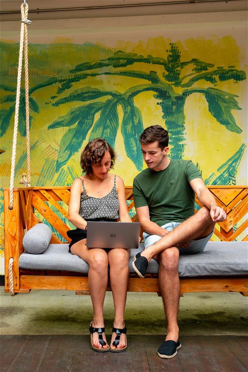 Woman and man sitting on a hanging wooden bench meeting with a laptop in front of a yellow and green painted wall at Selina