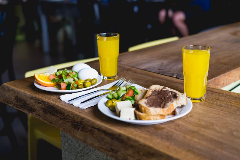 Two white plates on a wooden table of typical Israeli style breakfast including orange juice, toast, olives, Israeli salad, hard boiled eggs, fruit and cheese in Tel Aviv, Israel.