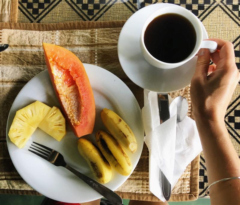 A white plate with pineapple, papaya and three tiny bananas and a hand with coffee at a hotel in Ella, Sri Lanka.