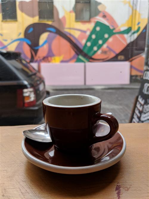 Brown coffee cup on a brown saucer with a silver spoon on a table overlooking a graffiti wall in Dublin, Ireland.