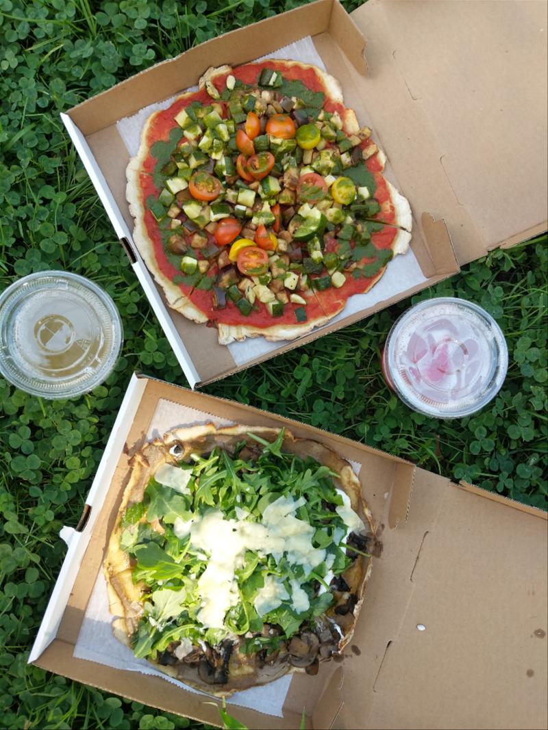 Two pizzas in brown cardboard carry-out boxes with cocktails to go on grass in a NYC park.